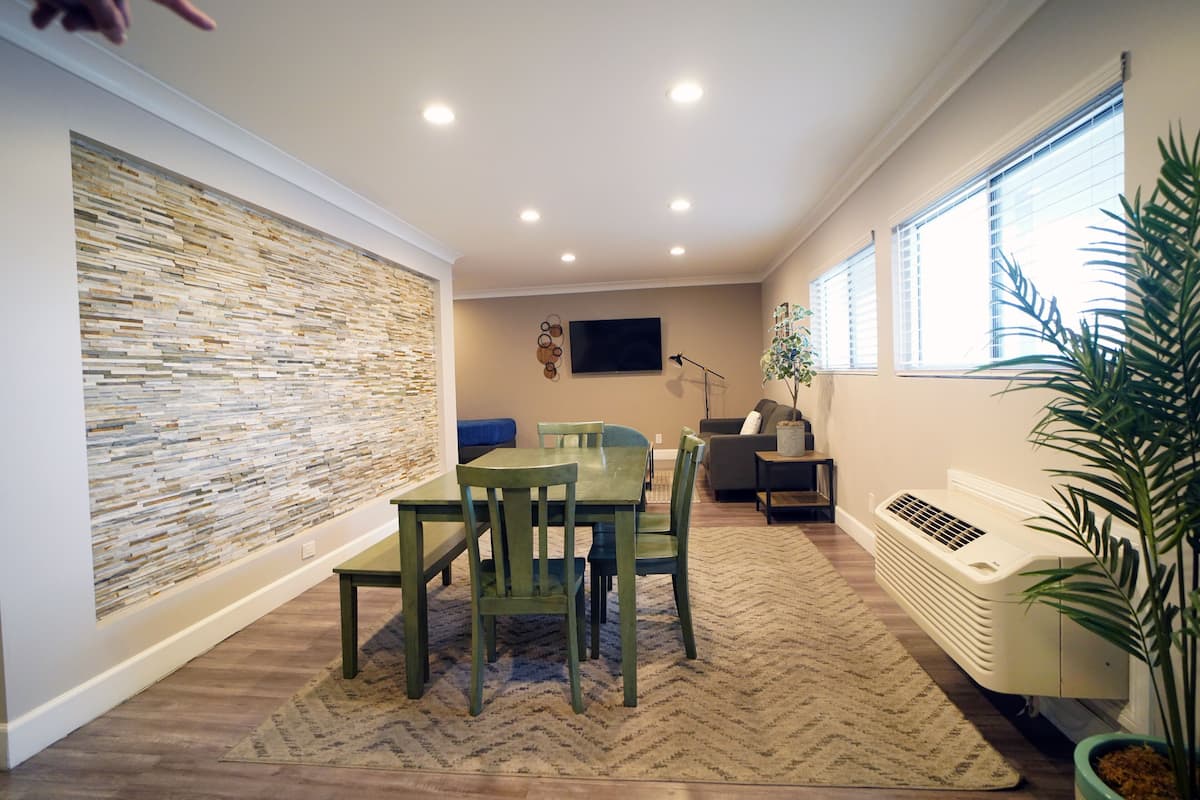 A modern living room with a stone wall, a dining table with chairs, a sofa, a TV, and a large window with blinds.
