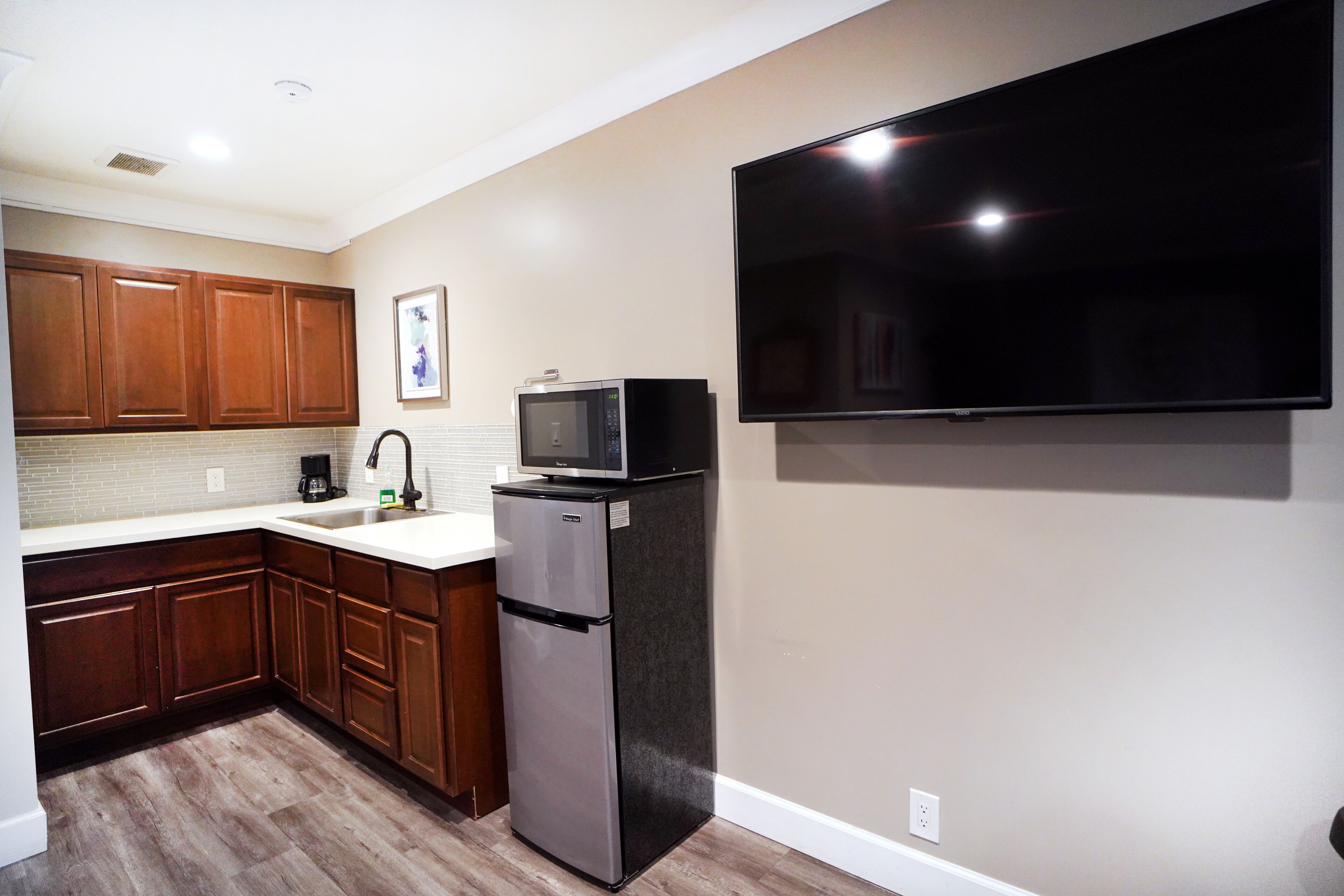A modern kitchen with wooden cabinets, a stainless steel refrigerator, a microwave, and a flat-screen TV mounted on the wall.