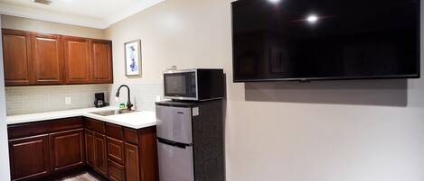 A modern kitchen with wooden cabinets, a stainless steel refrigerator, a microwave, and a flat-screen TV mounted on the wall.