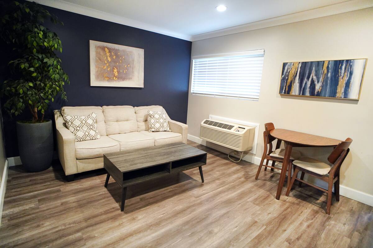 A living room with a beige sofa, a wooden coffee table, a dining table with chairs, a potted plant, and two wall-mounted artworks.