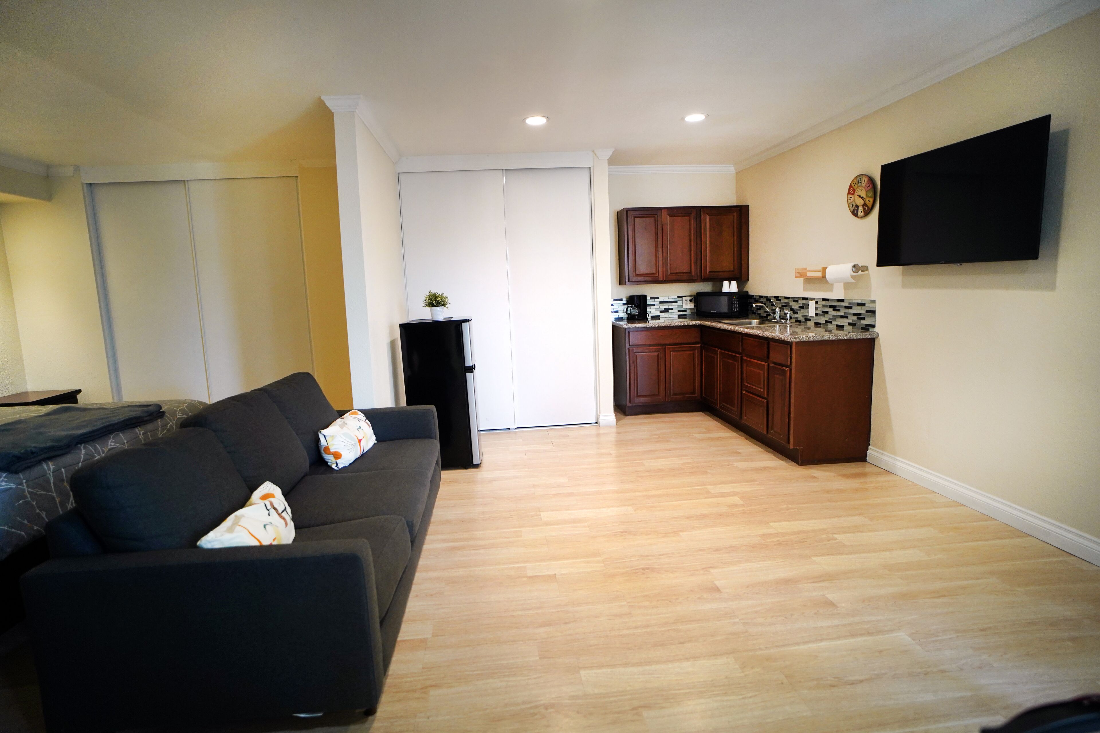 A living room with a dark-colored sofa, a kitchen area with wooden cabinets and a tiled backsplash, and a flat-screen TV mounted on the wall.