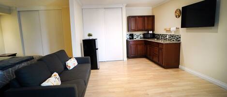 A living room with a dark-colored sofa, a kitchen area with wooden cabinets and a tiled backsplash, and a flat-screen TV mounted on the wall.