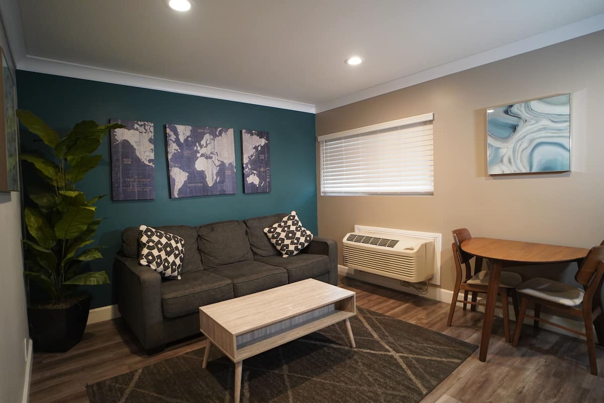A living room with a grey sofa, a wooden coffee table, a dining table with chairs, a large potted plant, and three world map paintings on the wall.