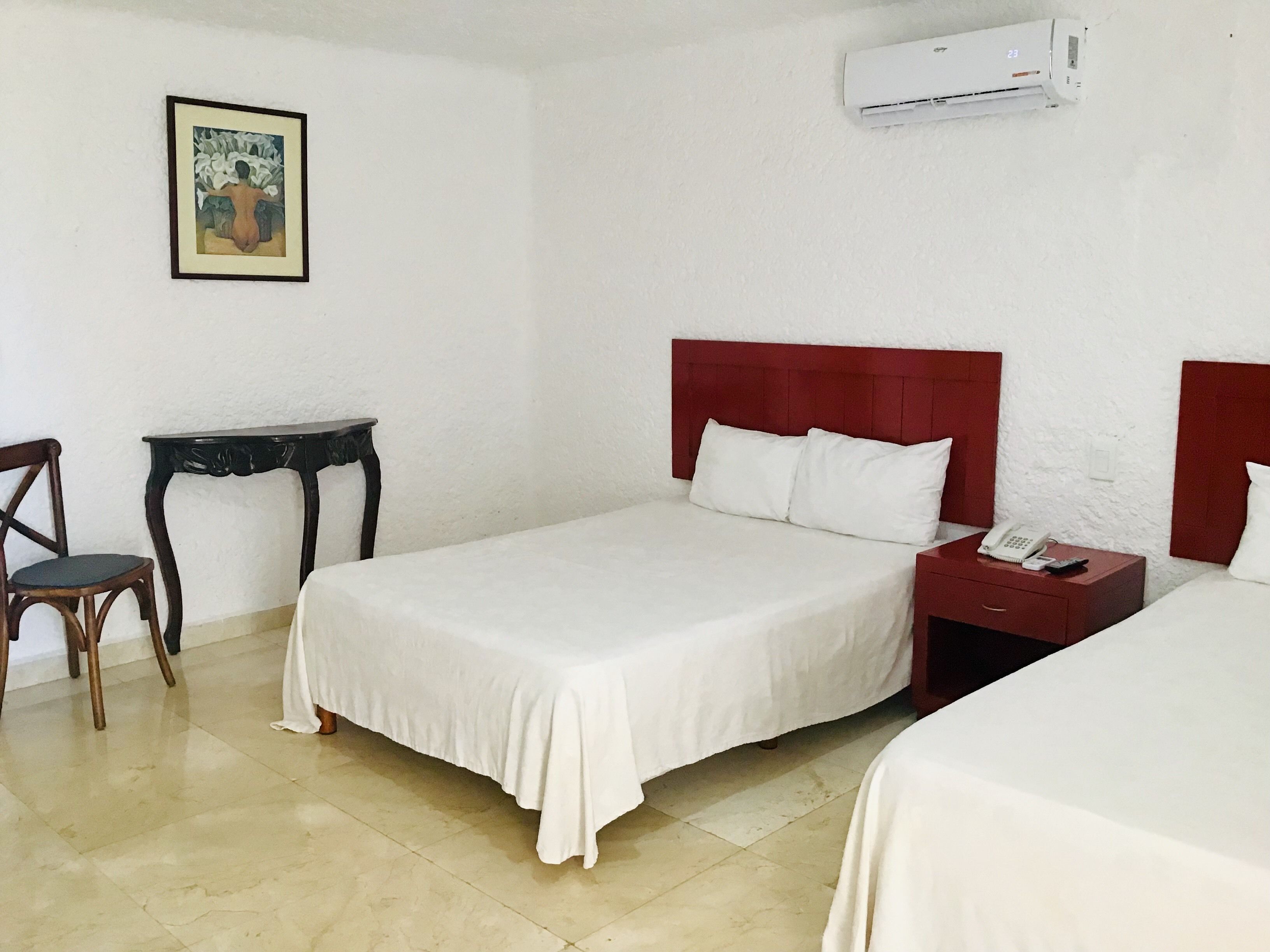 A hotel room with a bed, a chair, a small table, a bedside table with a phone, and an air conditioning unit.