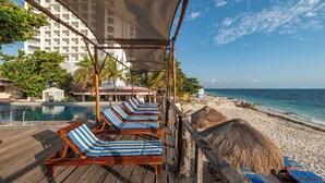A wooden deck with lounge chairs and a pool, overlooking a beach with a thatched hut and a hotel in the background. - Hotel Maya Caribe Faranda (Cancun)