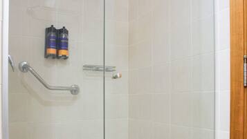 A glass-enclosed shower stall with white tiles, a grab bar, and two bottles of shampoo on the wall.