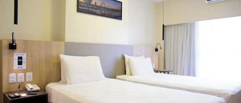 A hotel room with two beds, a nightstand with a phone and remote controls, a painting on the wall, and an air conditioning unit.