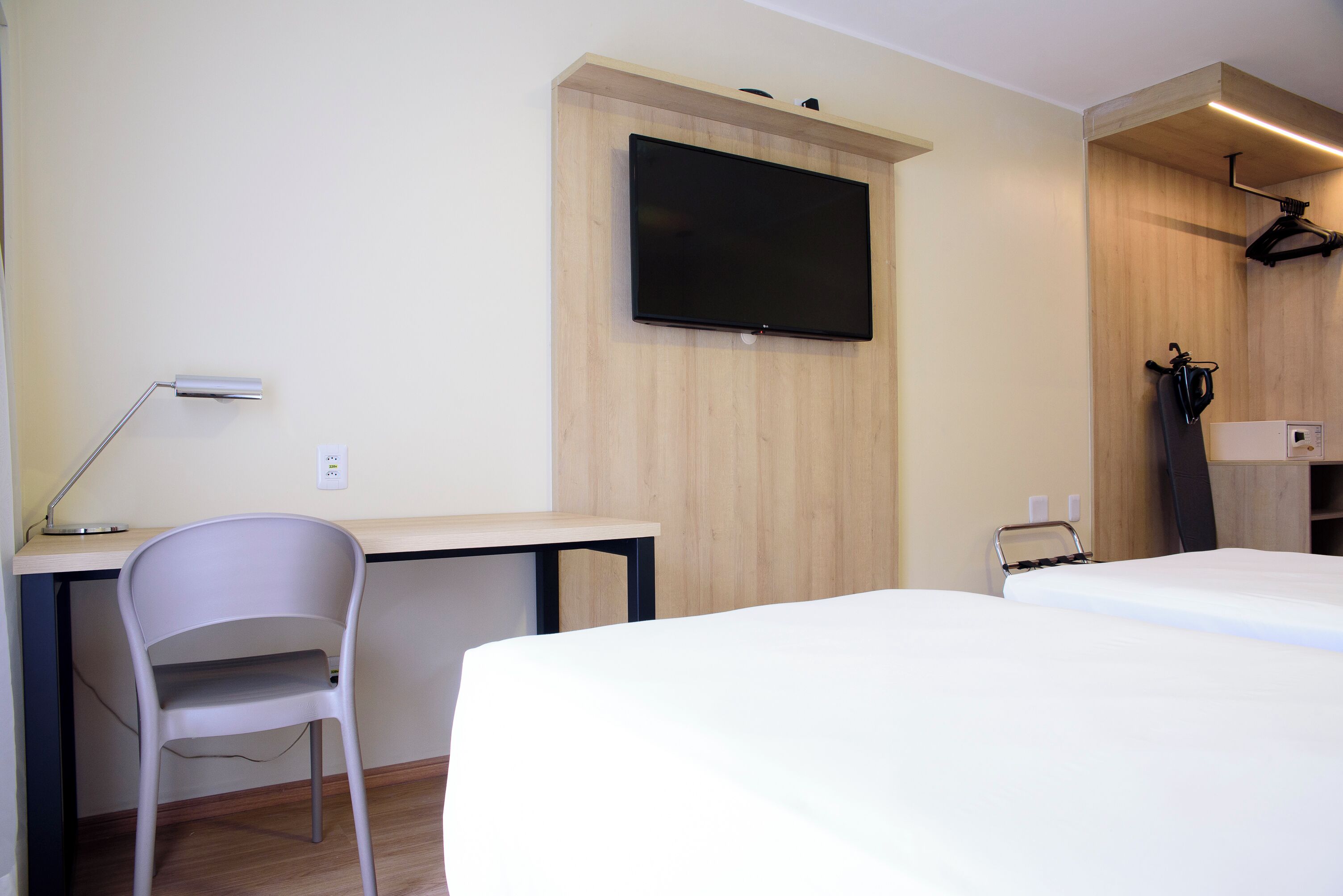 Ett hotellrum med en säng, ett skrivbord, en stol, en tv som är monterad på väggen och en klädställning.
