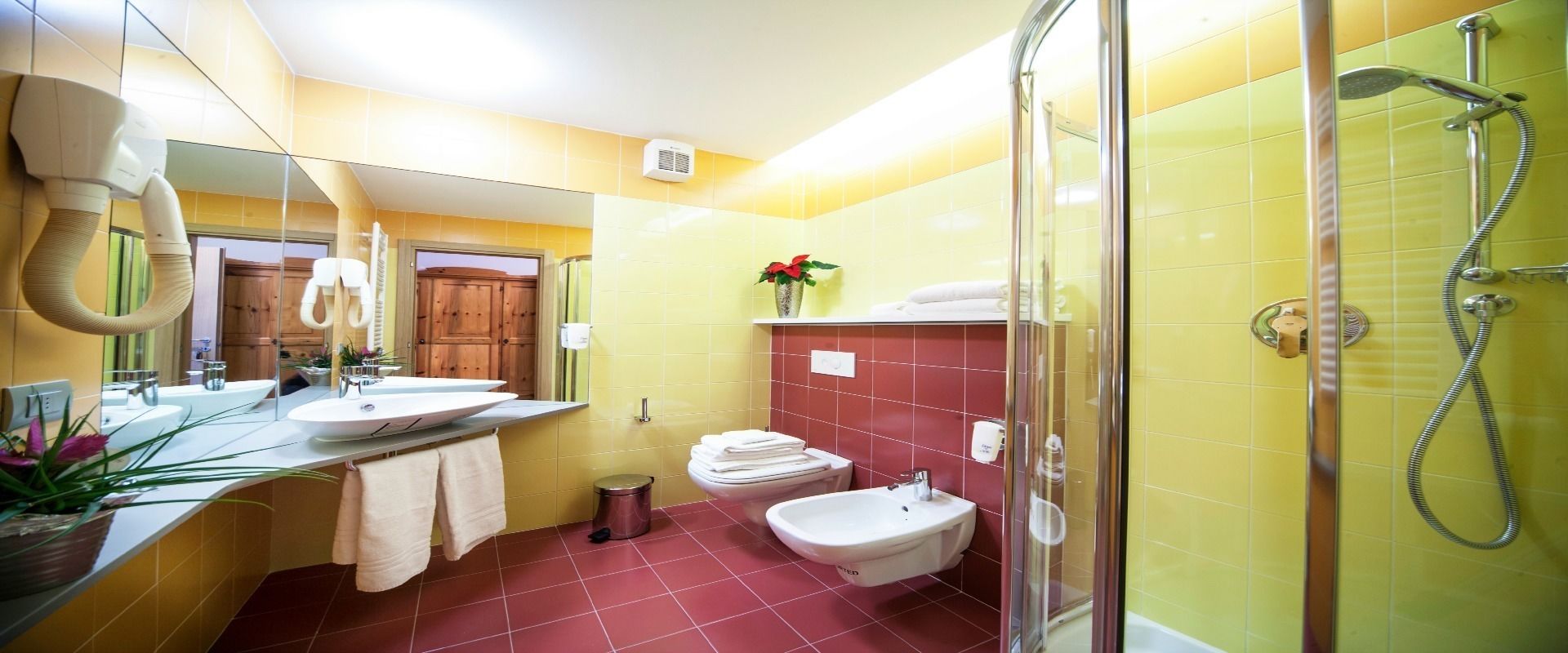 Un baño con baldosas amarillas y rojas, ducha, bidé, inodoro y lavamanos con espejo.