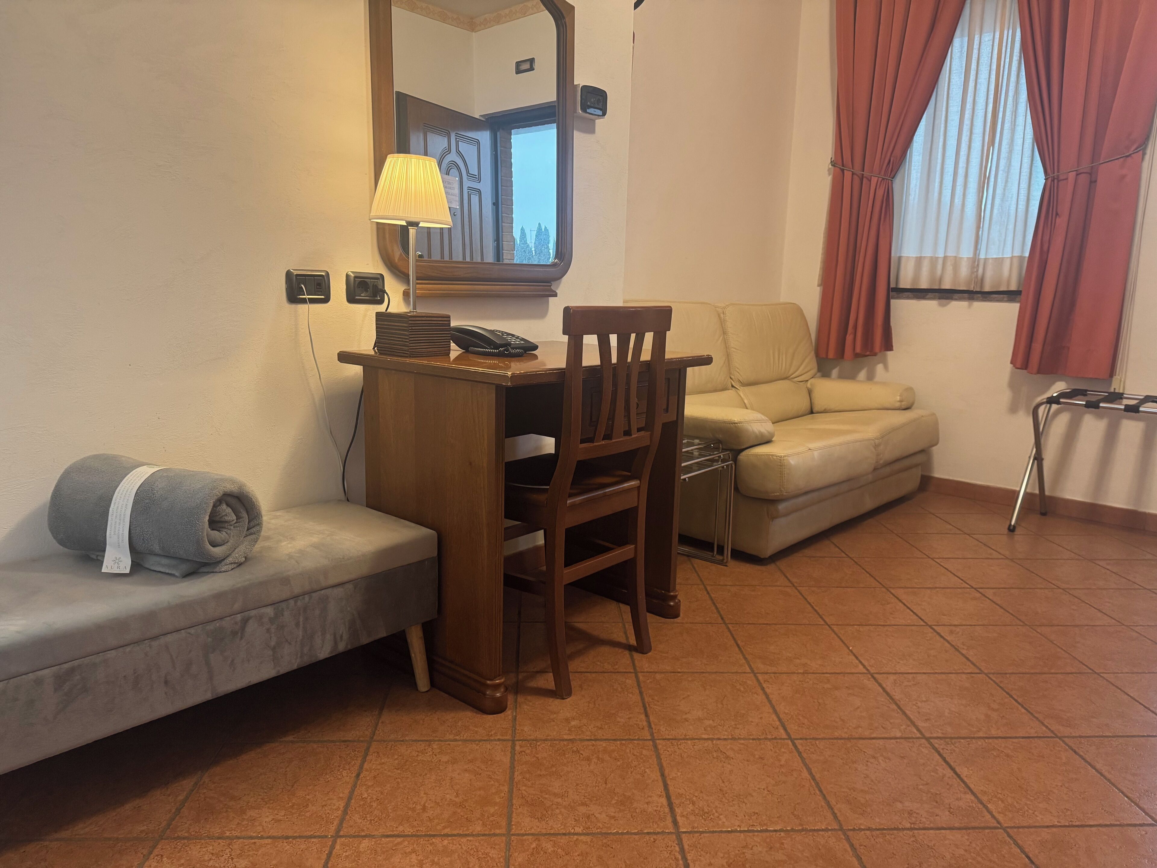 Een kamer met een houten bureau, een zachte bank, een sofa, een spiegel, een lamp en een raam met gordijnen.