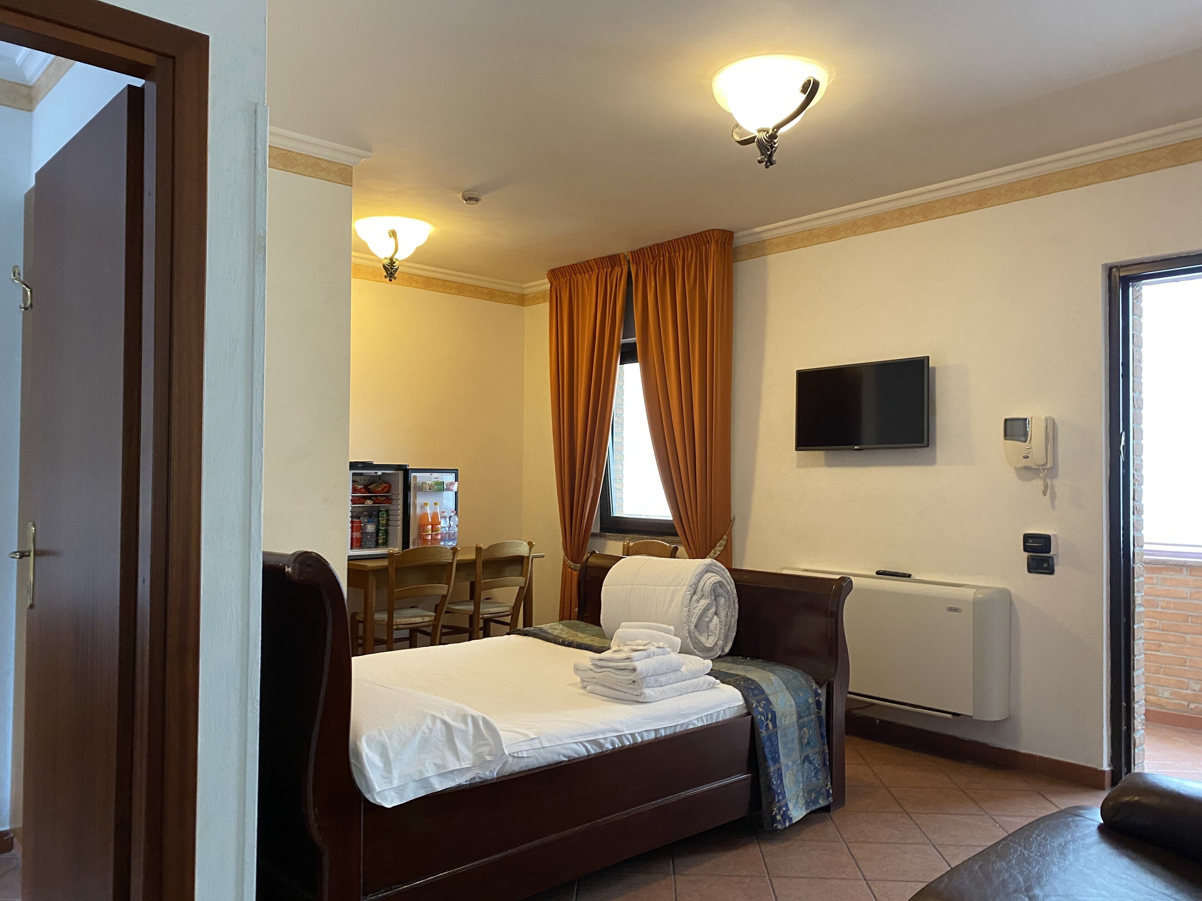 Quarto de hotel com cama, escrivaninha com refrigerador, televisão e janela com cortinas.