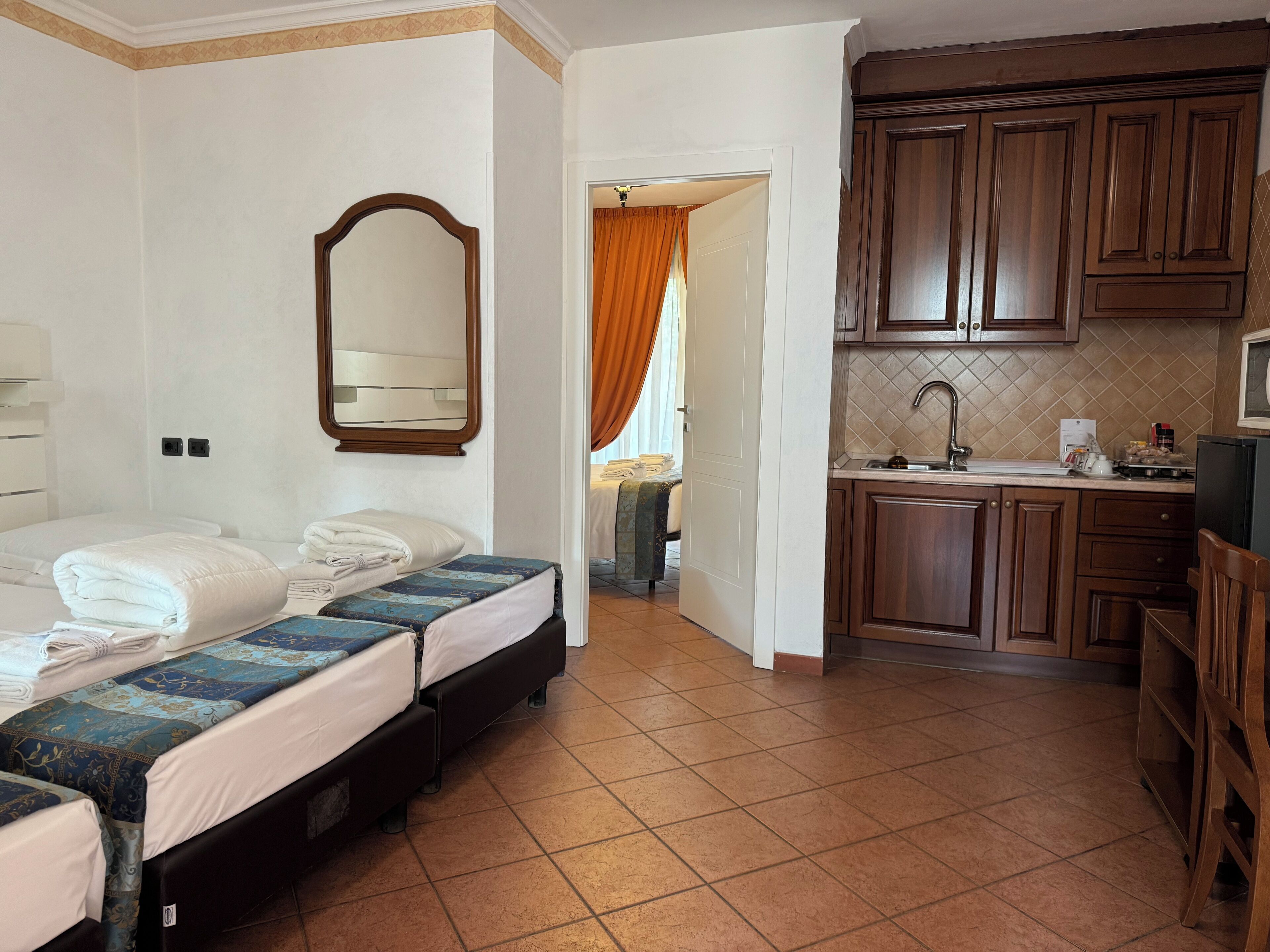 Habitación de hotel con dos camas, un espejo grande, una cocina con armarios de madera y piso de baldosas.