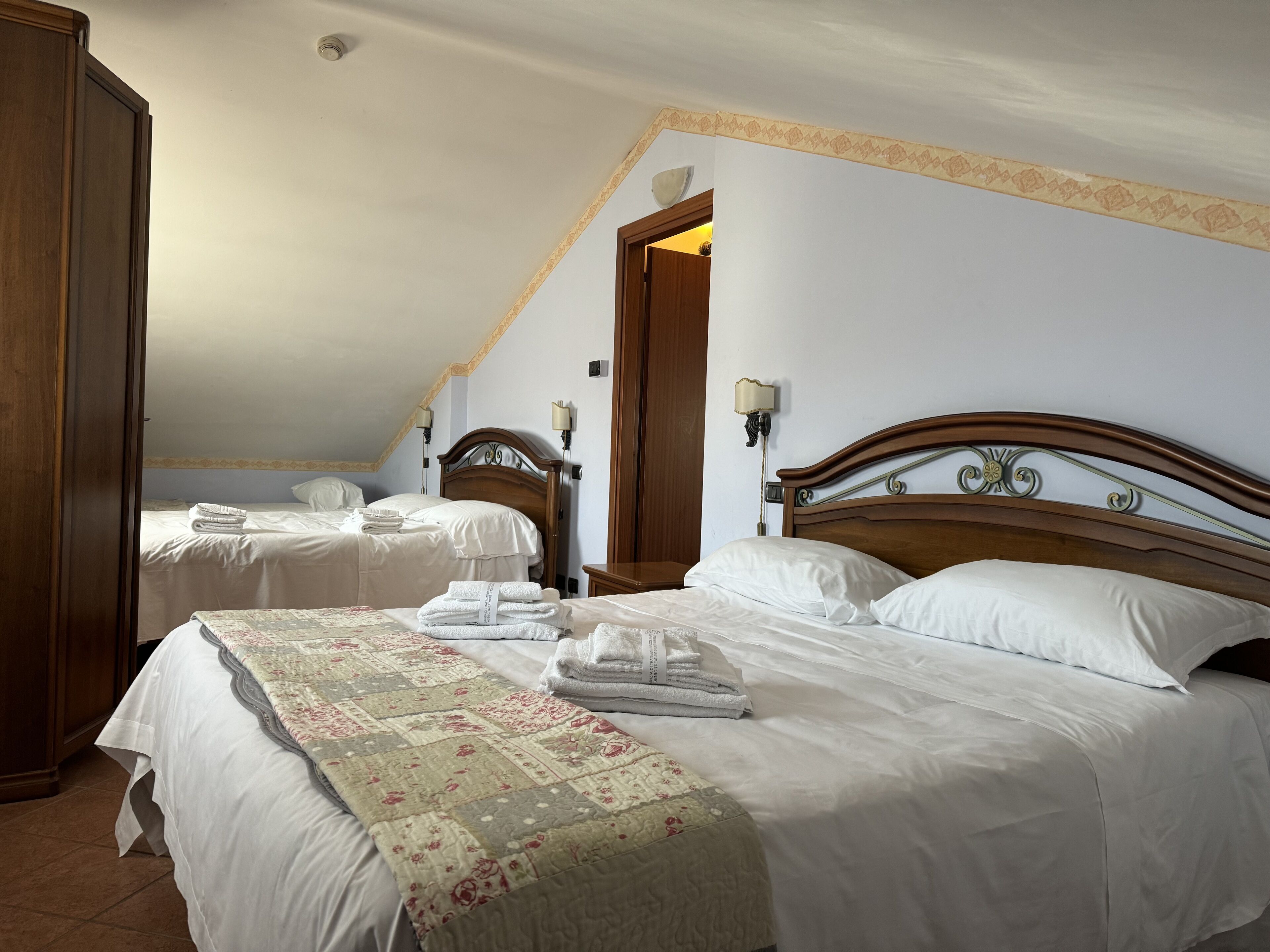 A hotel room with two beds, each with white linens and a floral-patterned bedspread. There are two wall-mounted lamps and a wooden wardrobe.