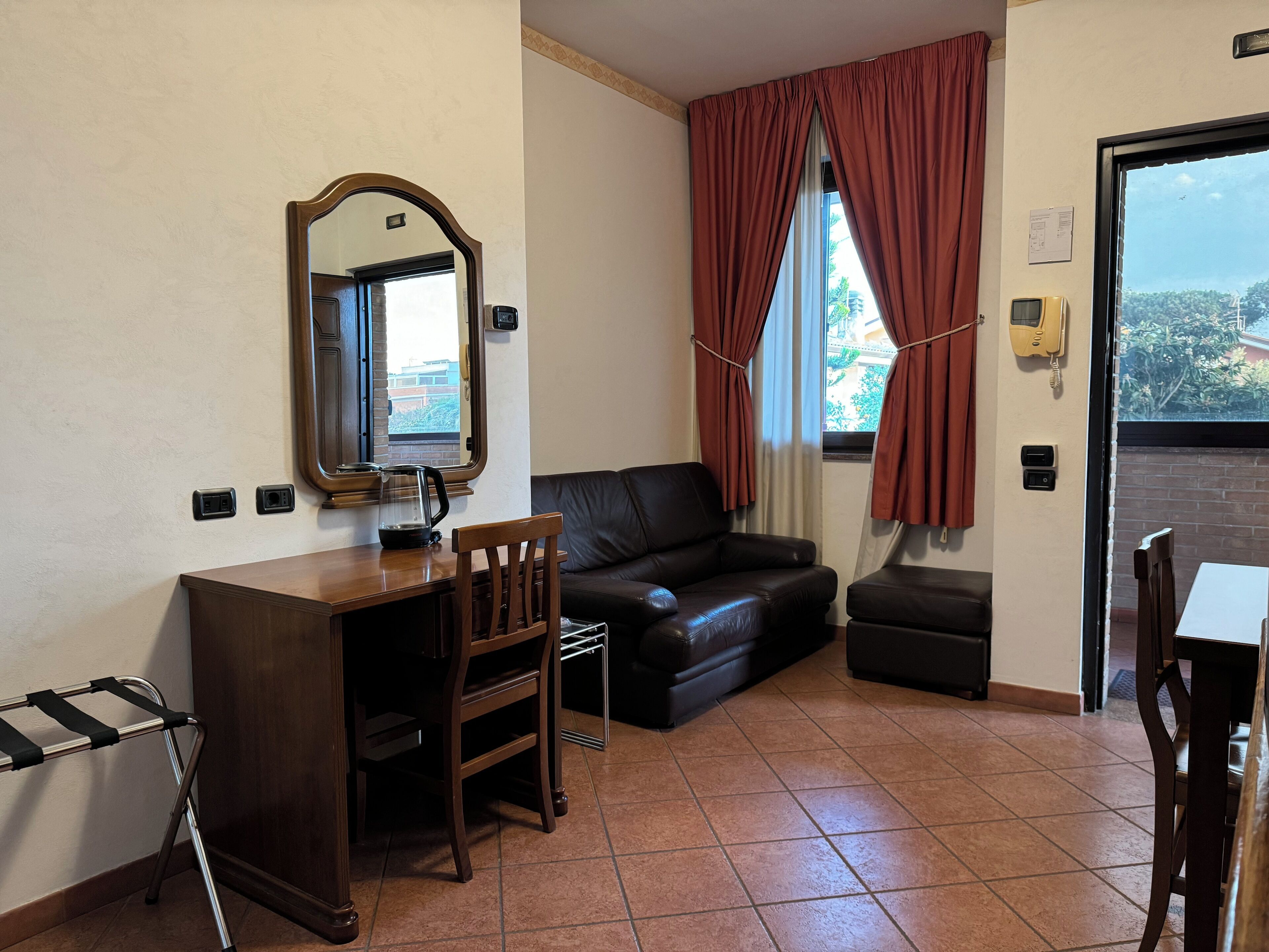 Ein Zimmer mit Schreibtisch aus Holz, einem Sessel, einer Ledersofa, einem kleinen Tisch und einem Fenster mit roten Vorhängen.