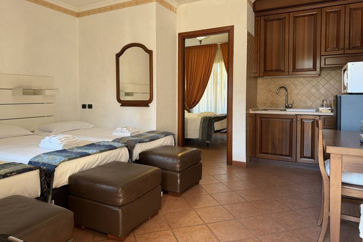 Habitación de hotel con cama, dos mesitas de noche, un espejo, una kitchenette pequeña y un baño visible a través de una puerta abierta.