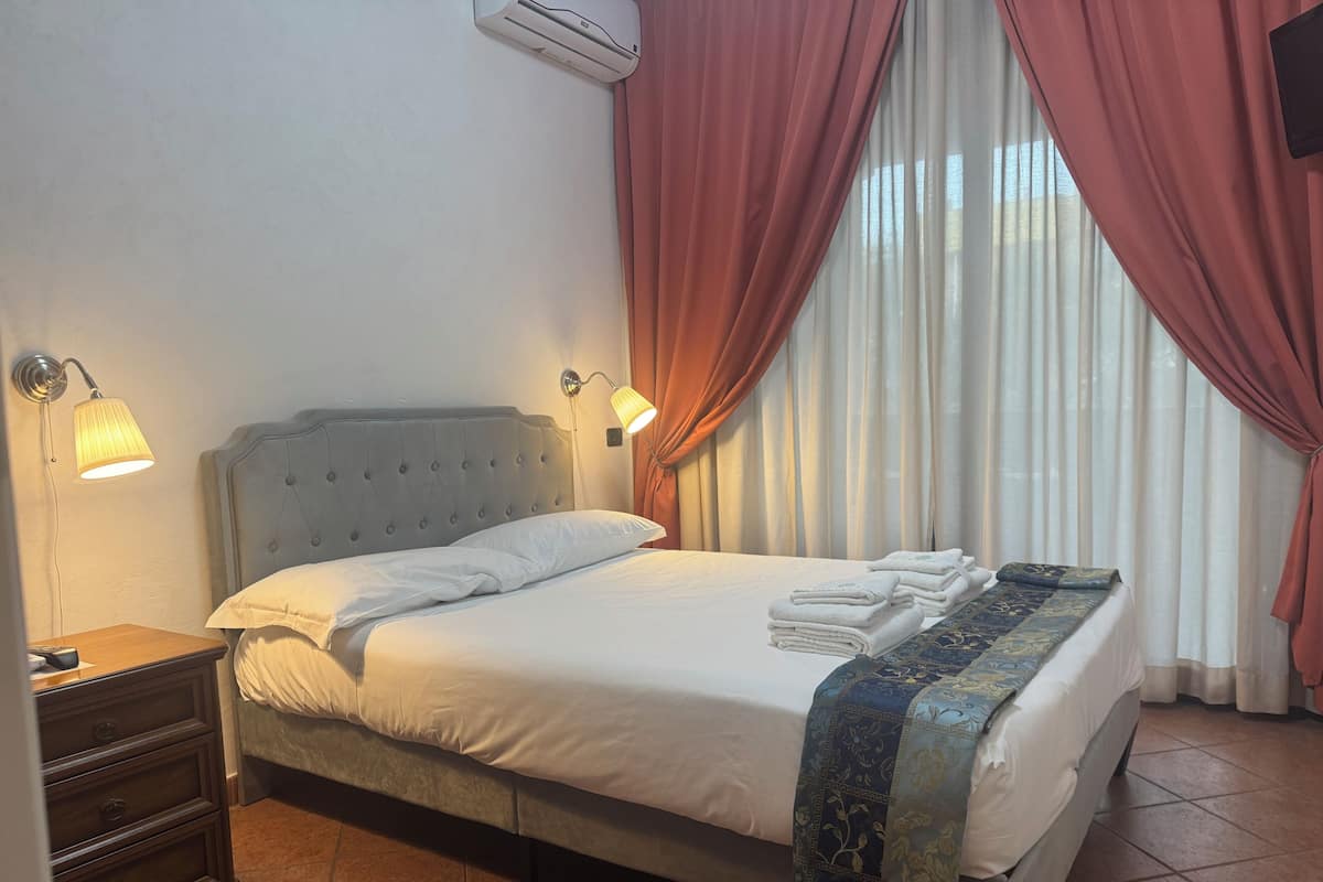 Una cama bien hecha con edredón blanco y una manta decorativa azul, una lámpara de noche y una mesita de noche con un cajón de madera.