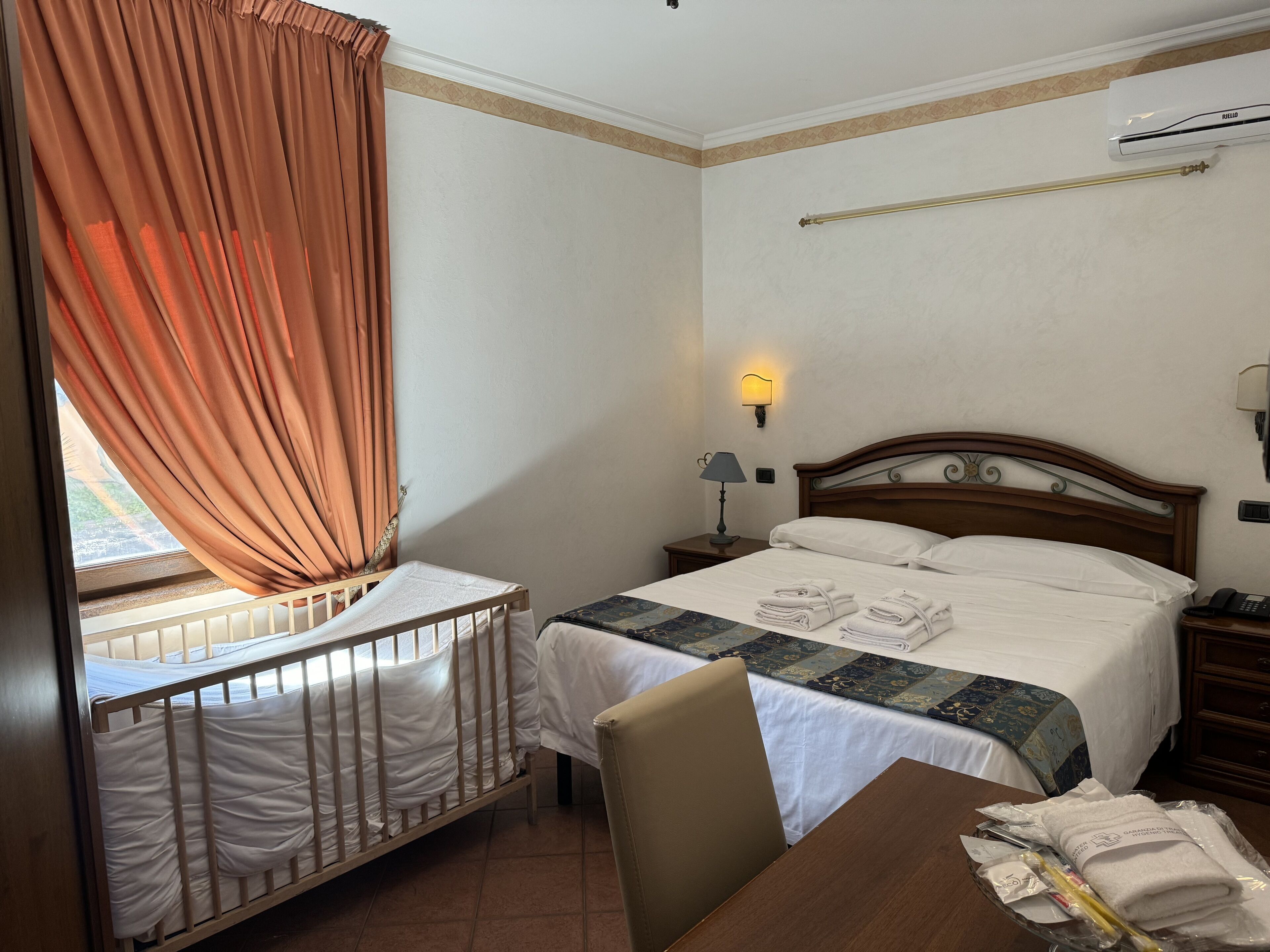 A hotel room with a crib, a bed, a desk, and a window with curtains.