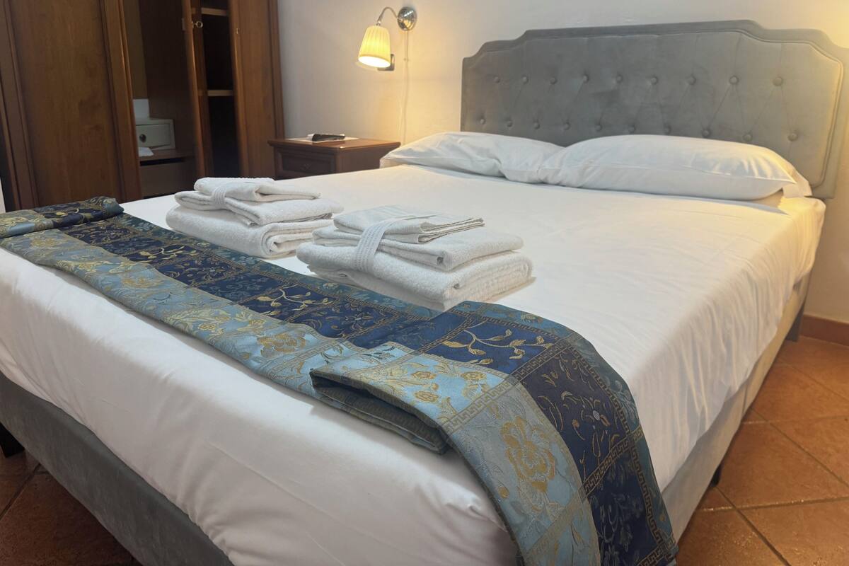 Una cama bien hecha con un edredón estampado, una toalla blanca doblada y una lámpara de noche.