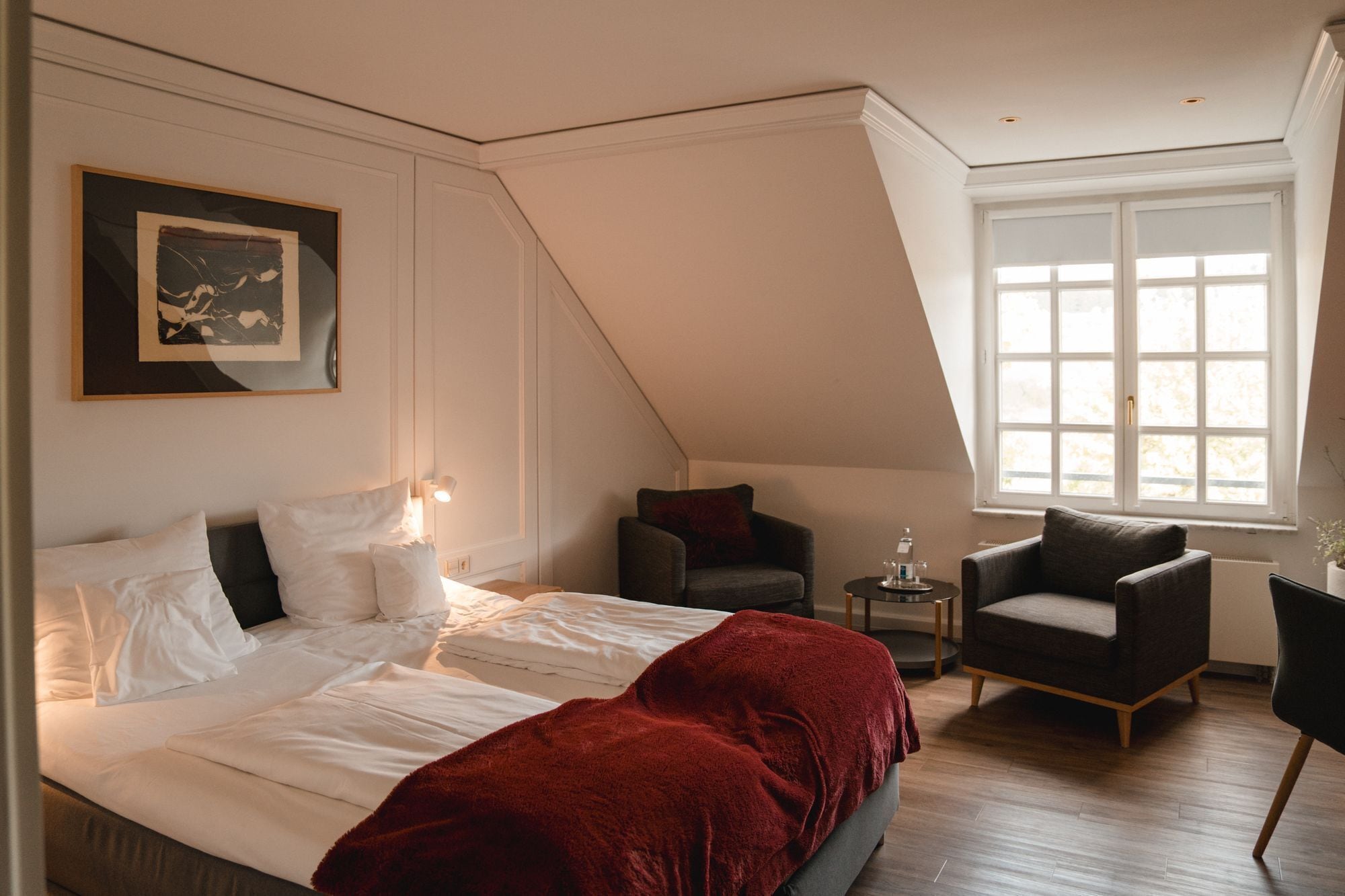 Un dormitorio con una cama grande, una manta roja, una silla, una mesita de noche con una lámpara y una ventana con persianas.