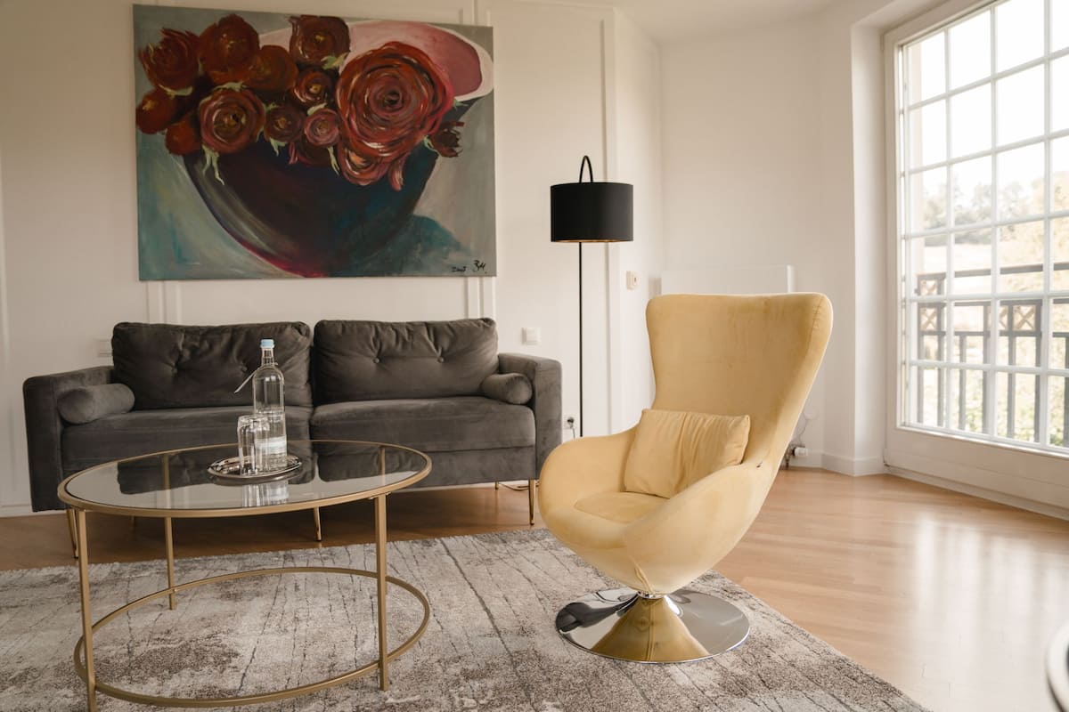 A living room with a grey sofa, a round glass coffee table, and a yellow armchair. There is a painting of flowers on the wall and a floor lamp.