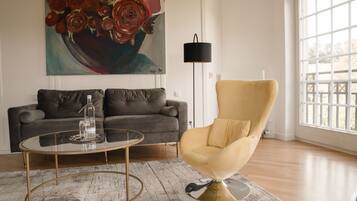 A living room with a grey sofa, a round glass coffee table, and a yellow armchair. There is a painting of flowers on the wall and a floor lamp.