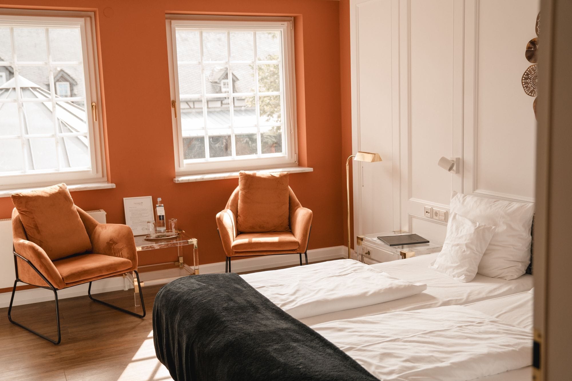 Une chambre avec deux chaises, un lit, une table de chevet avec une lampe et une fenêtre donnant sur des immeubles.