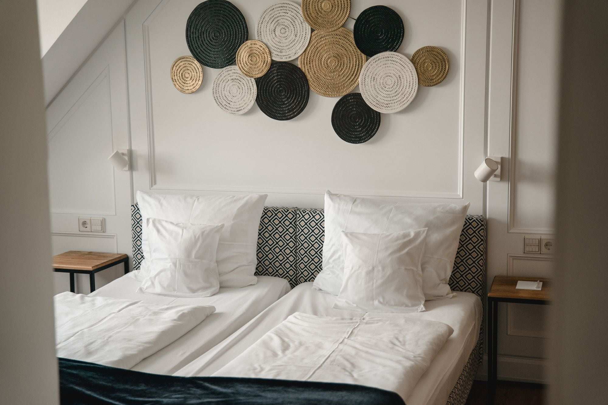 Ein ordentlich bezogenes Bett mit weißen Leinen, einem Kissenbezug mit geometrischem Muster und einem an der Wand hängenden gewebten Kunstwerk darüber.