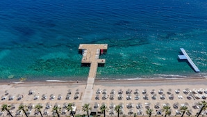 An aerial view of a beach with a pier extending into the sea, rows of beach umbrellas, and palm trees. - Latanya Park Resort - All Inclusive (Bodrum)