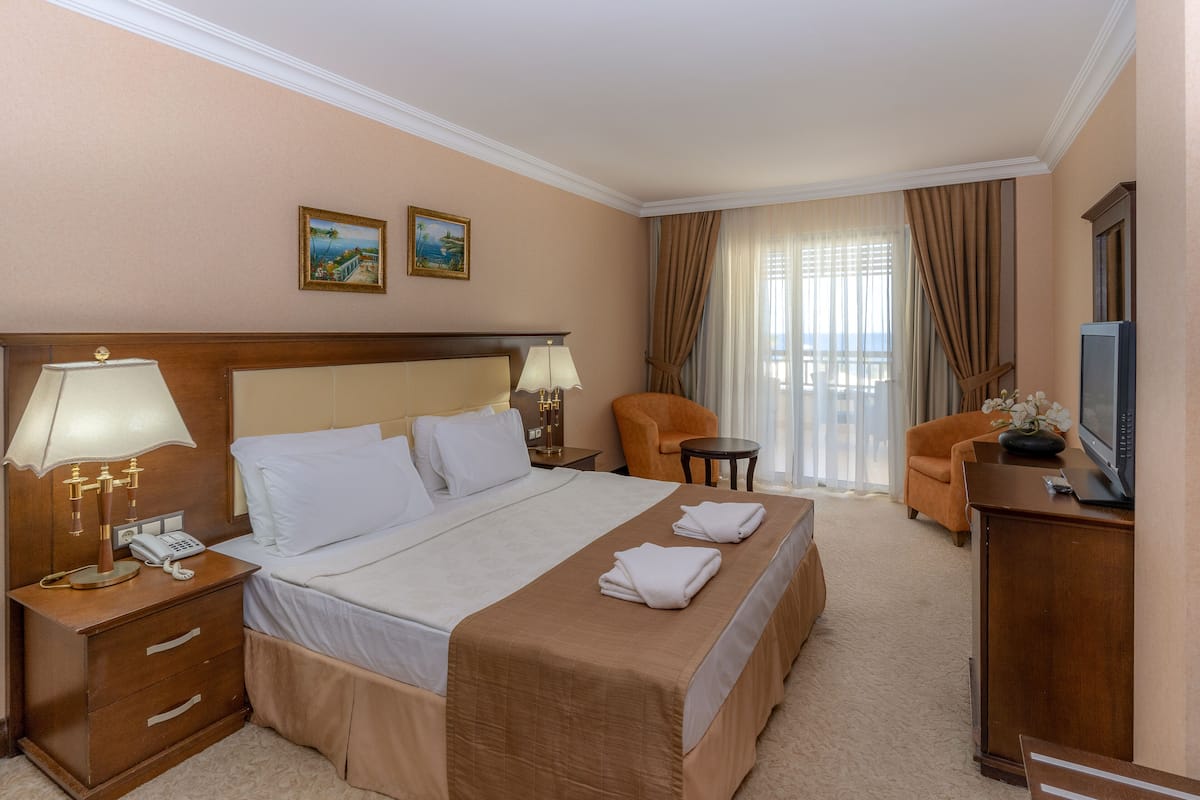 Habitación de hotel con una cama grande, dos mesitas de noche, una silla, un escritorio con un televisor y un ventanal con cortinas.