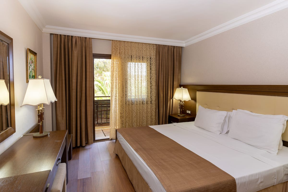 Habitación de hotel con una cama grande, dos lámparas de noche, una mesita de noche de madera, un espejo y una puerta que da a un balcón.