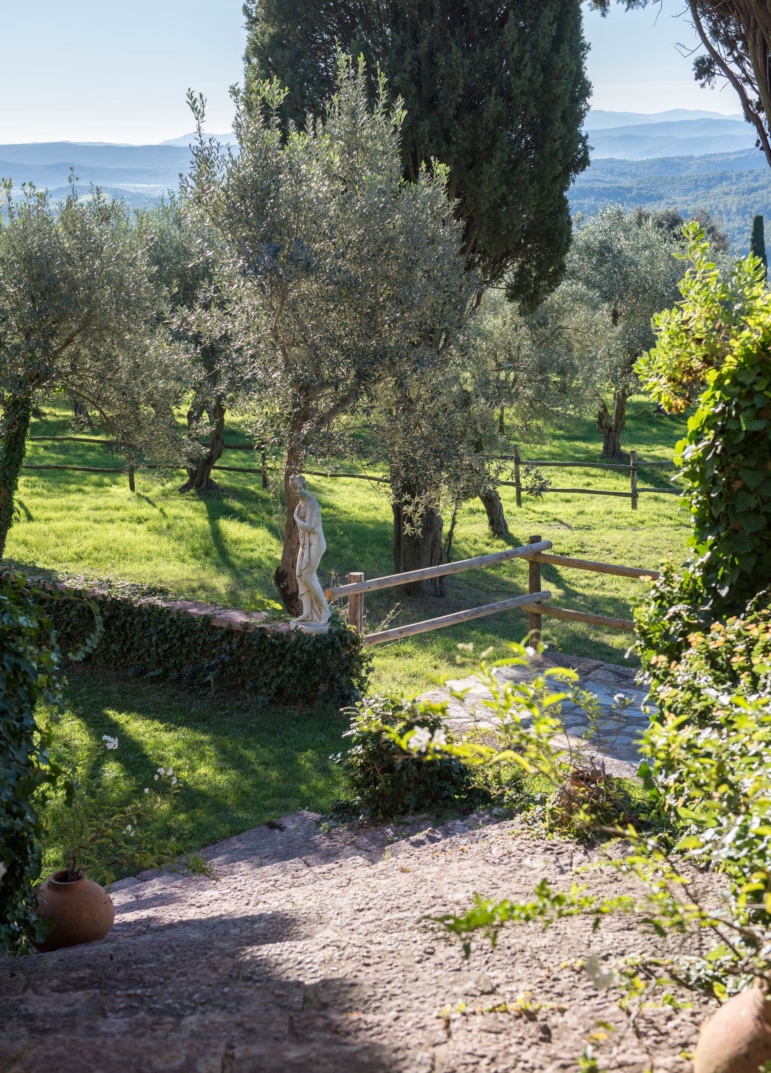 Un sentier de pierres rustiques traverse un jardin luxuriant, parsemé d’oliviers et bordé d’une clôture en bois.