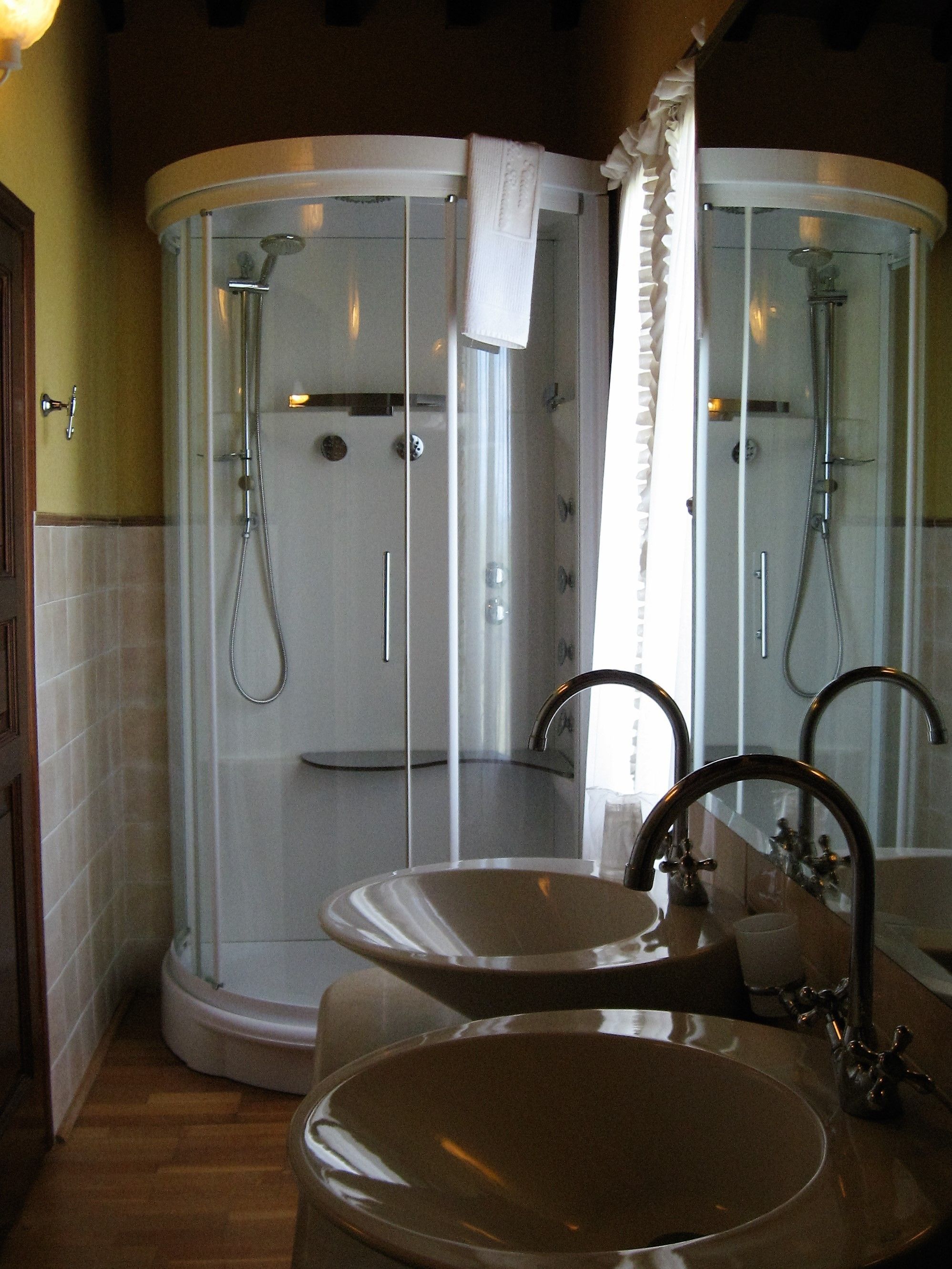 Un baño con ducha envuelta en vidrio, dos lavamanos y un espejo.