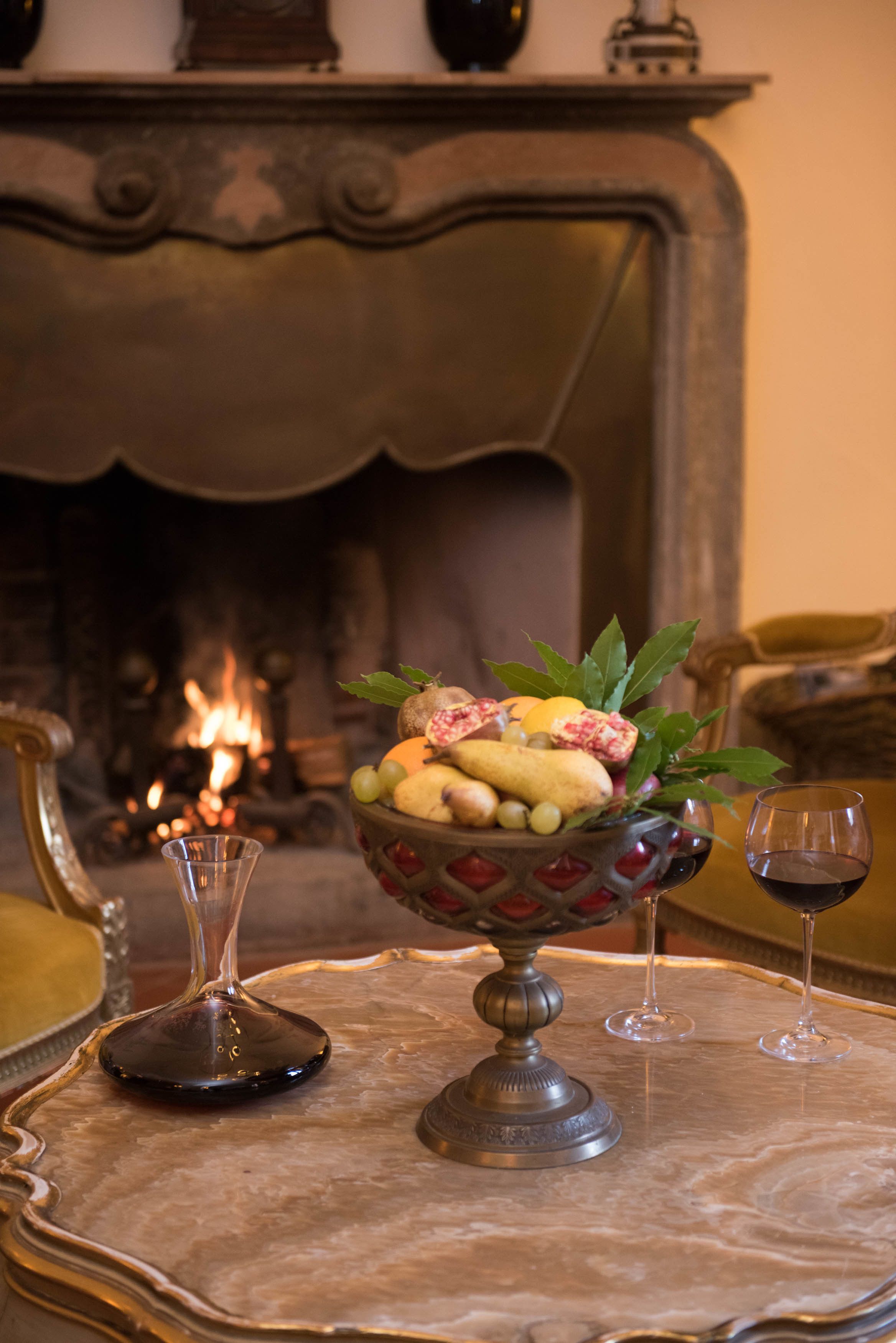 A table with a decorative bowl of fruit and two wine glasses, set against a fireplace backdrop.