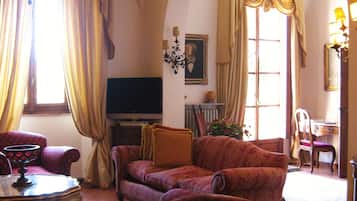 Uma sala de estar com um candelabro, sofás vermelhos estampados, uma televisão e um espaço de refeições com uma cadeira e mesa.