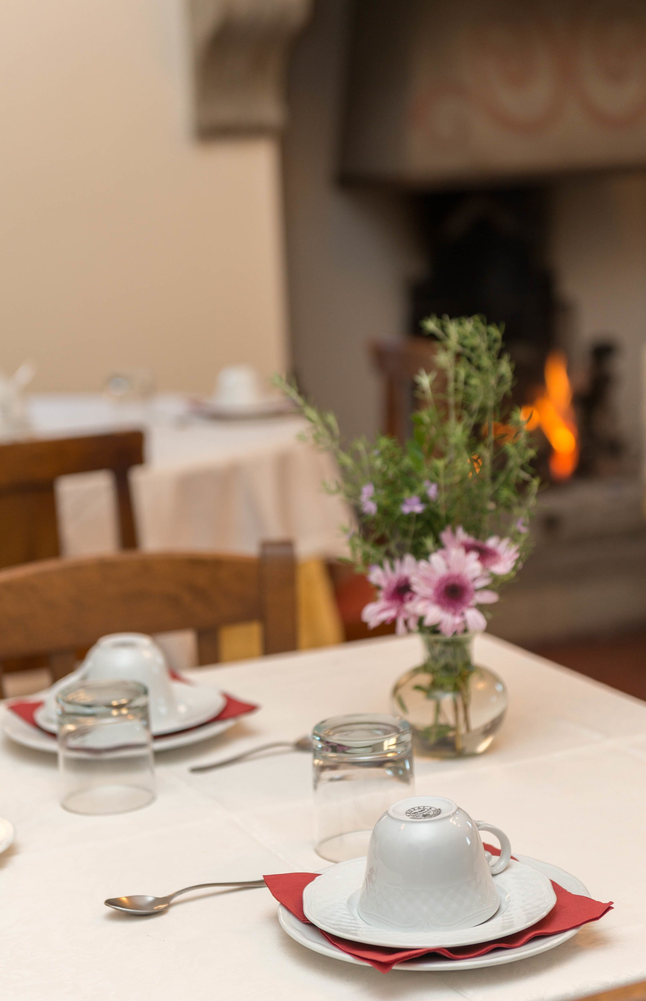 Une table à manger est dressée avec de l’assiette blanche, une tasse blanche et un verre d’eau. Un vase rempli de fleurs se trouve à l’arrière-plan.