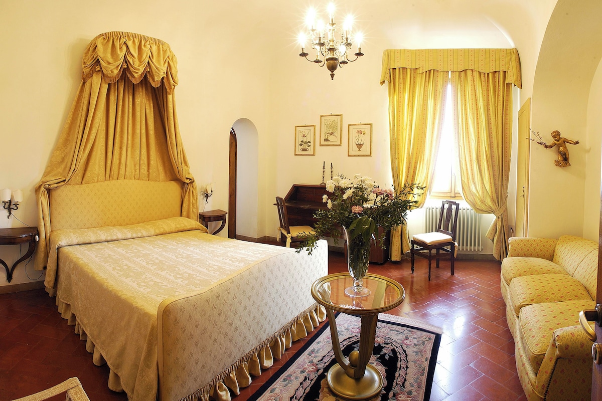 A room with a bed, a chair, a table with a vase of flowers, and a sofa.