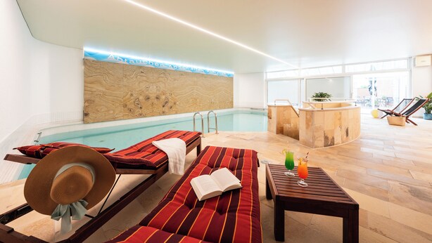 A poolside area with lounge chairs, a wooden wall with a lighted sign, a bar area, and a view of the outdoors. - Best Western Hotel Heide (Oldenburg)