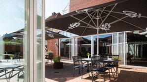 An outdoor patio area with tables and chairs under umbrellas, a brick floor, and a glass door leading inside. - Best Western Hotel Heide (Oldenburg)
