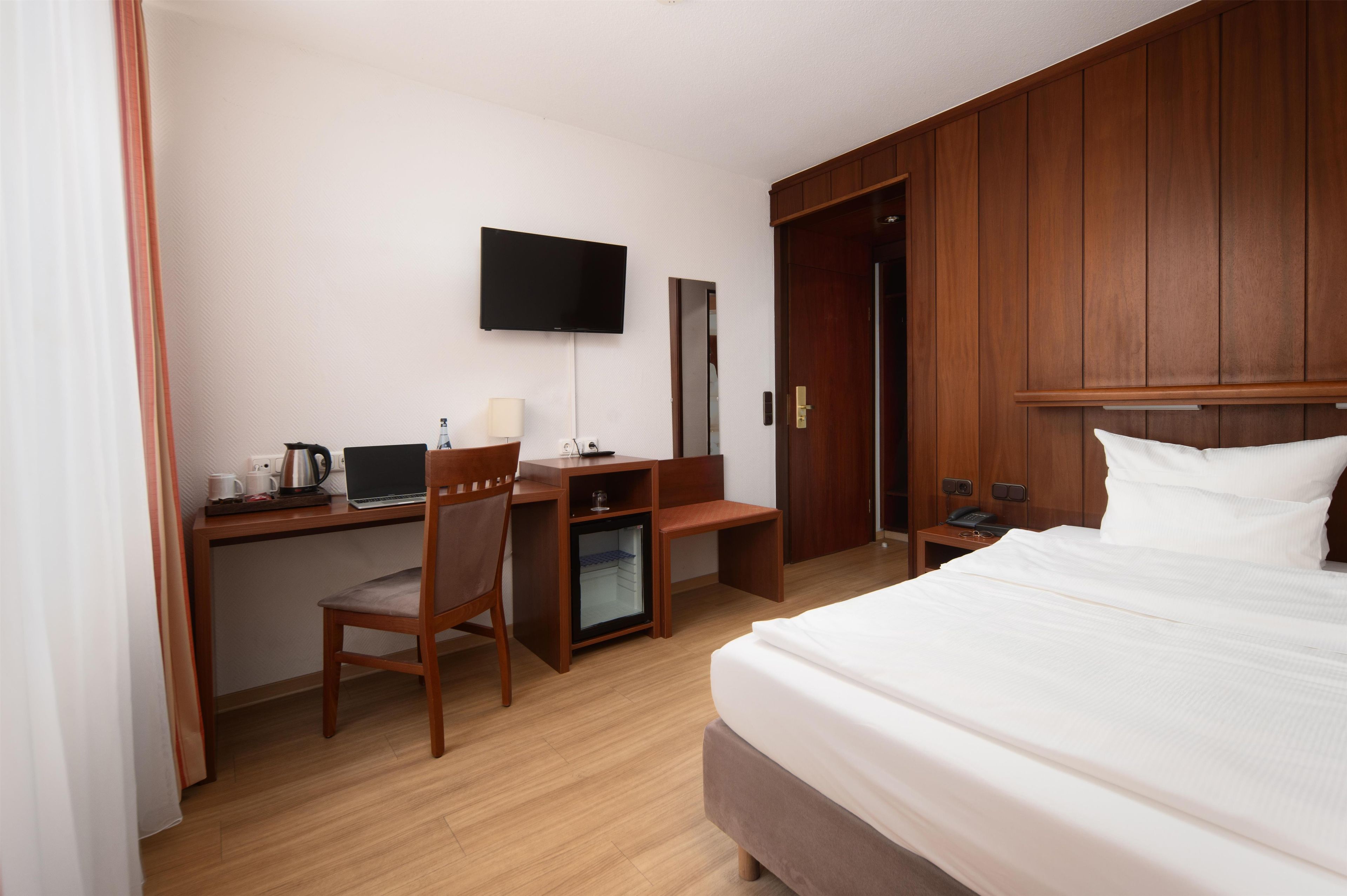 A hotel room with a bed, a desk with a computer, a chair, a small refrigerator, a TV mounted on the wall, and a wardrobe.