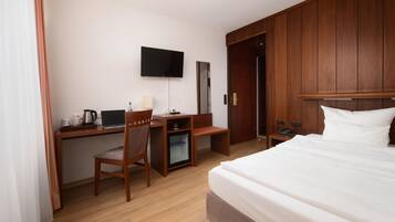 A hotel room with a bed, a desk with a computer, a chair, a small refrigerator, a TV mounted on the wall, and a wardrobe.