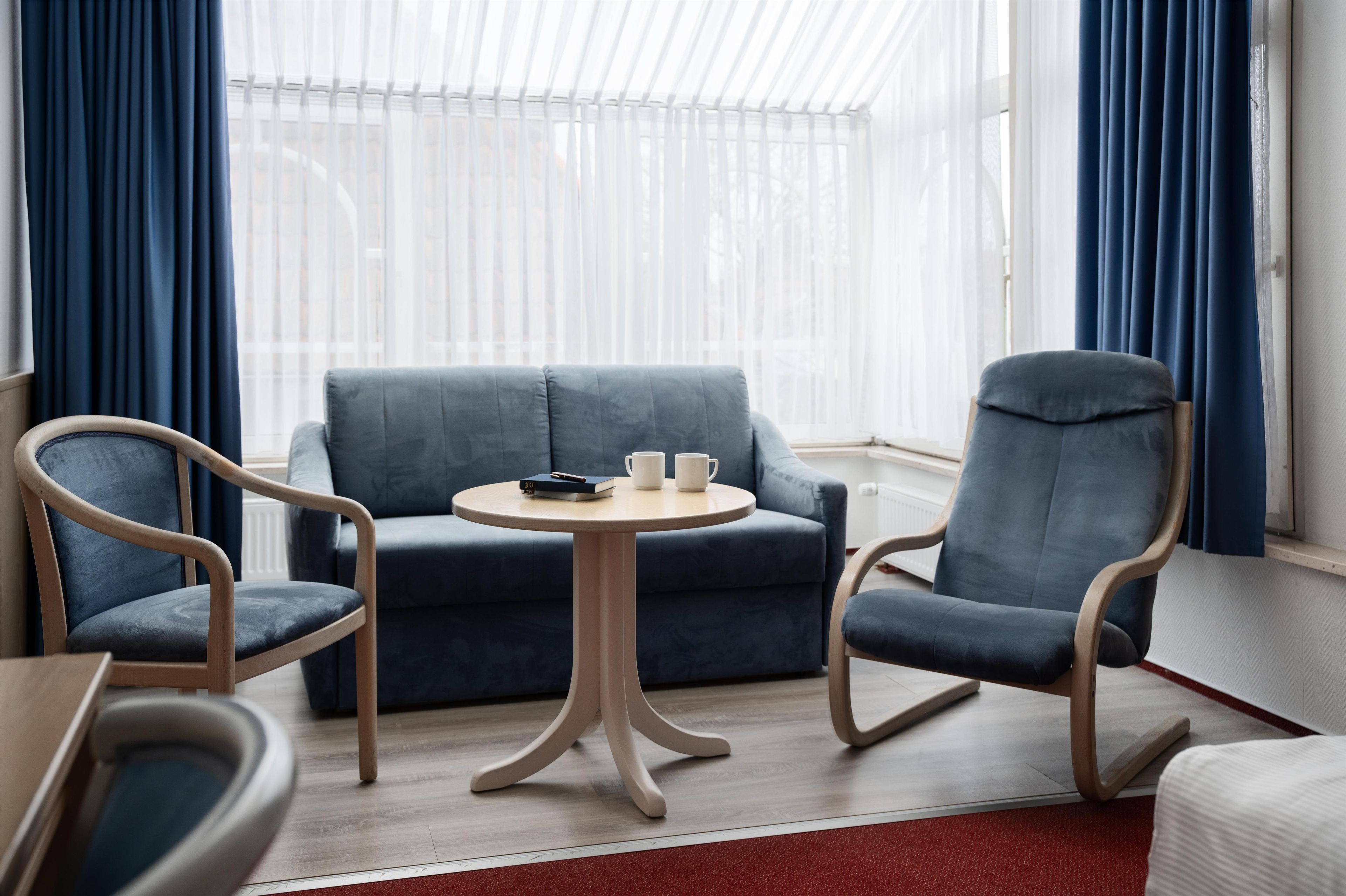 A room with a blue sofa, two wooden rocking chairs, a small round table with a book and a mug, and sheer white curtains.