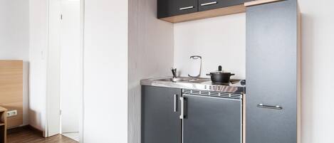 Une cuisine compacte équipée d’un four encastré, d’un évier et d’une cuisinière. Un meuble en bois se trouve au-dessus de la cuisinière, et une porte donne accès à une autre pièce.