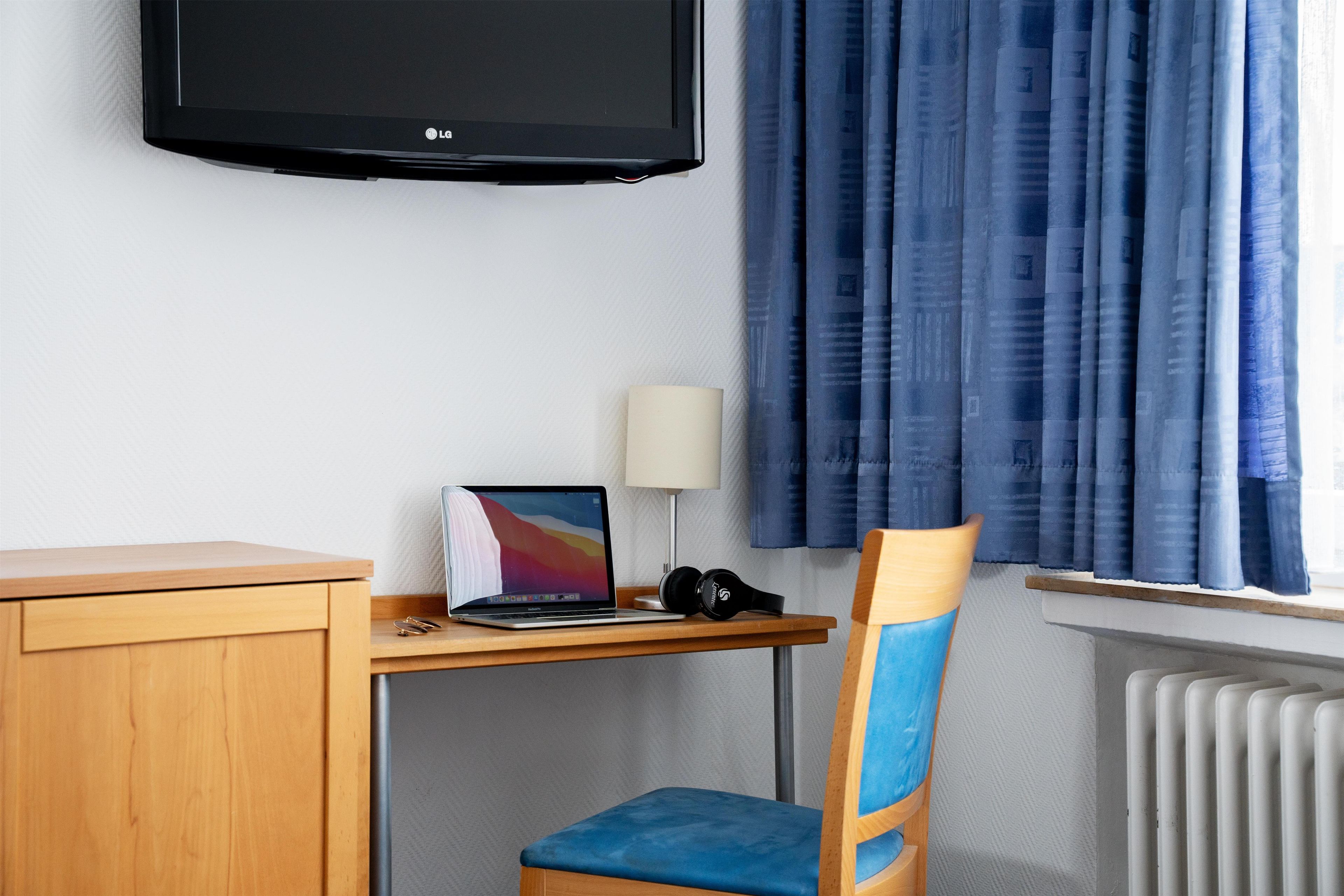 A hotel room with a desk, a laptop, a chair, a TV, a lamp, and a window with curtains.