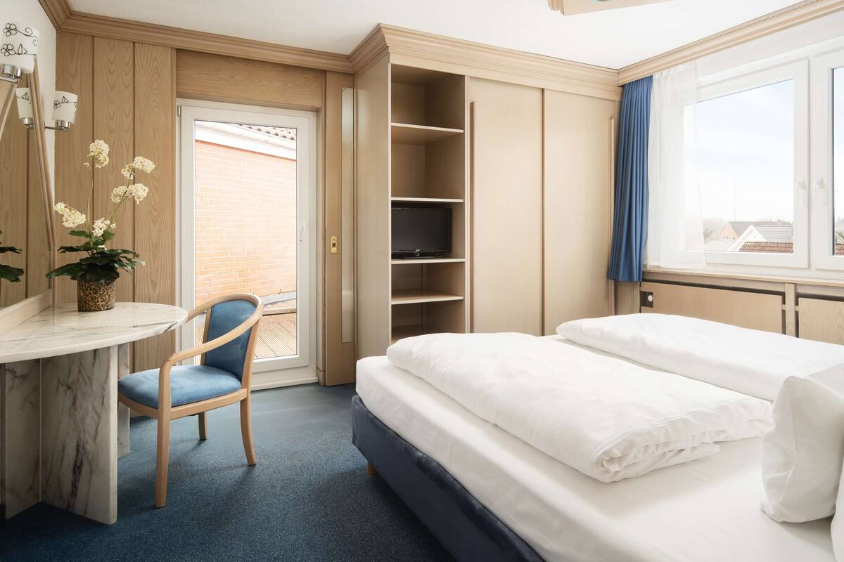 Una habitación de hotel con cama, escritorio, silla, televisor y ventana con cortinas.