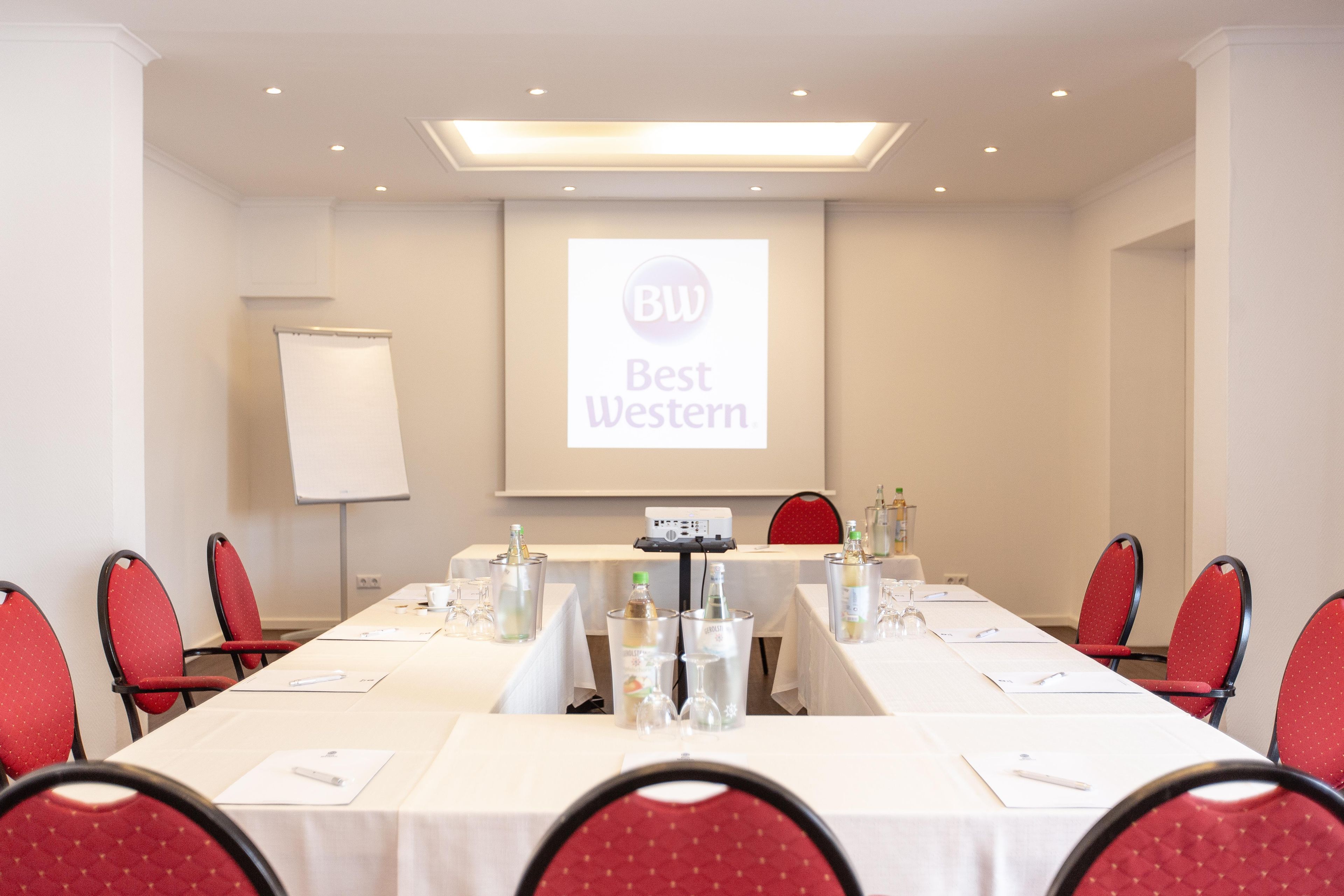 A conference room with a long table, red chairs, and a projector screen displaying 'Best Western' branding.