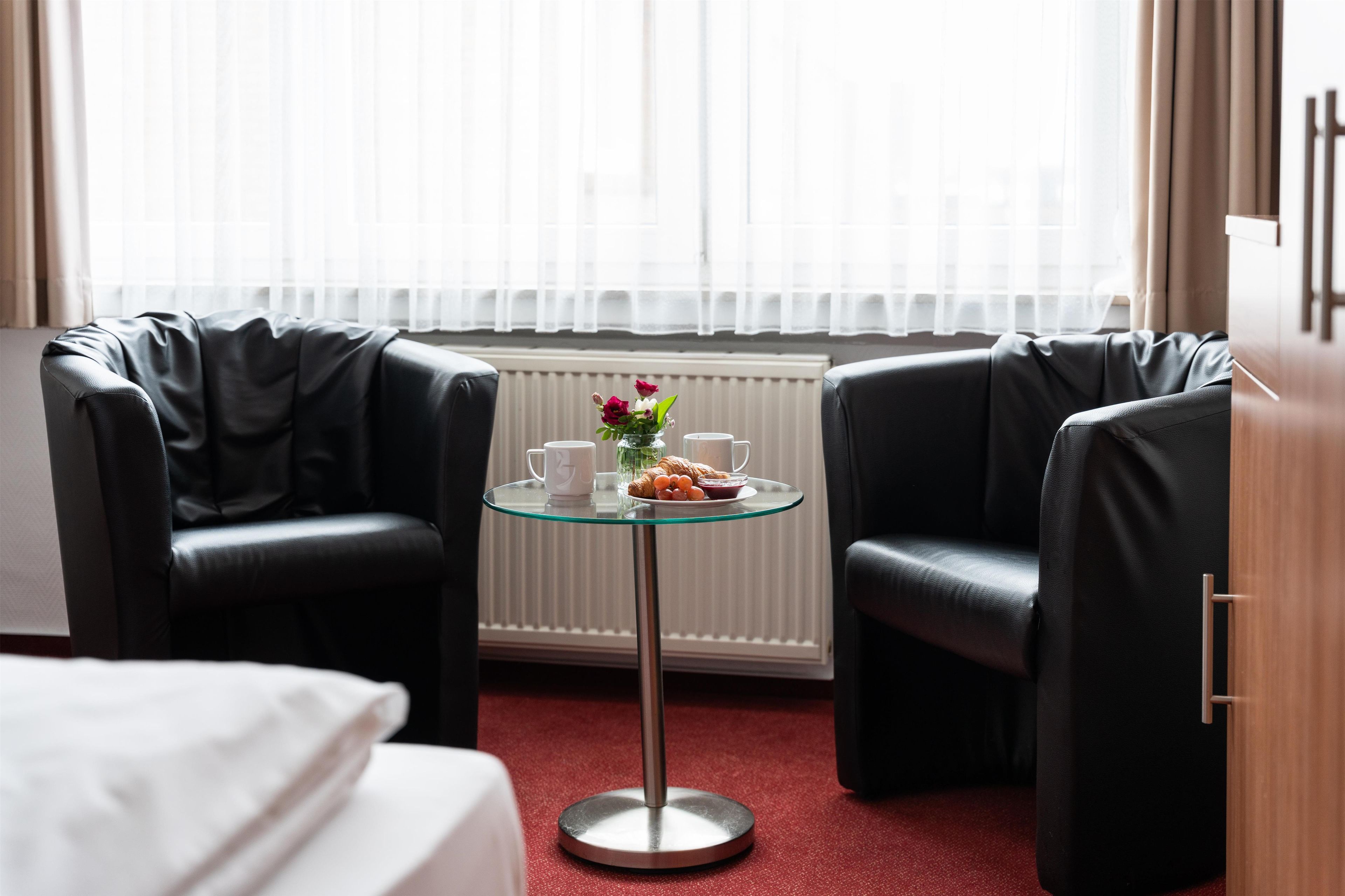 A hotel room with two black leather chairs, a glass table with a white mug, a plate of food, and a vase with flowers.