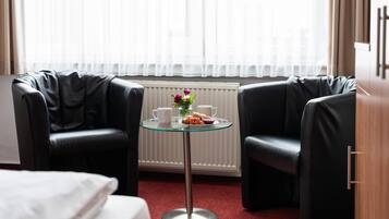 A hotel room with two black leather chairs, a glass table with a white mug, a plate of food, and a vase with flowers.