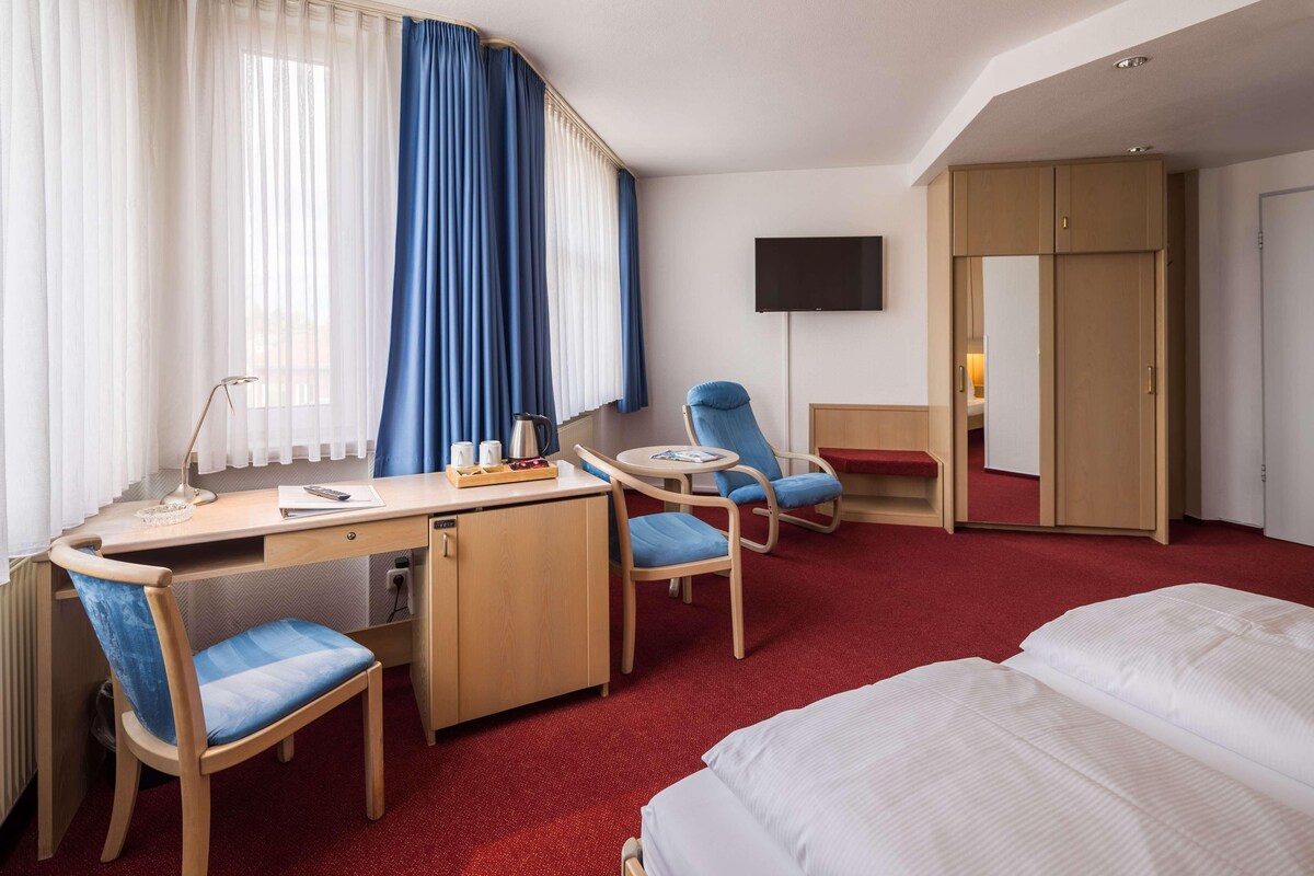 A hotel room with a bed, desk, chairs, a TV, and a wardrobe.