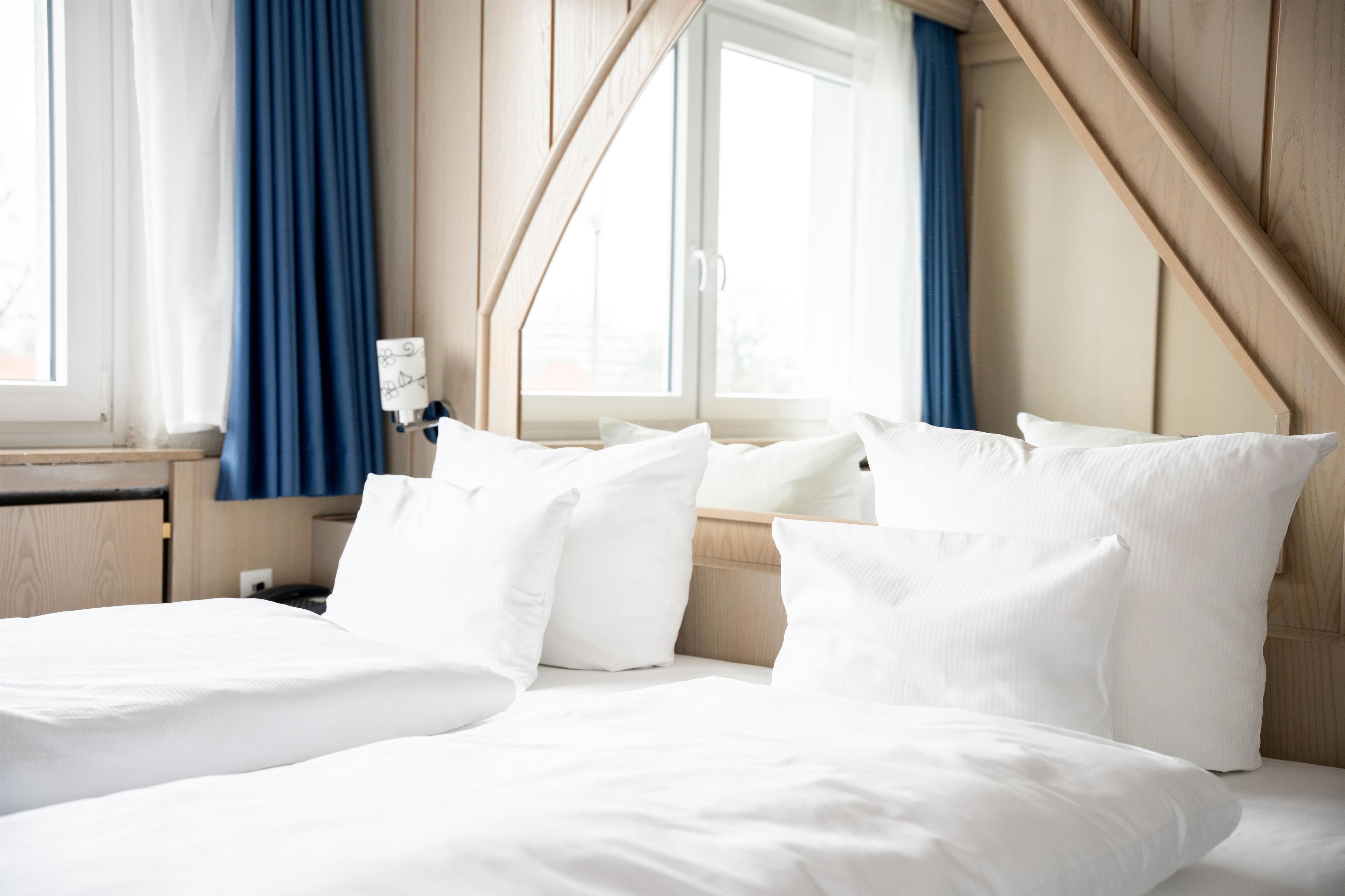 Habitación de hotel con cama, almohadas, mesita de noche, lámpara y ventana con cortinas.