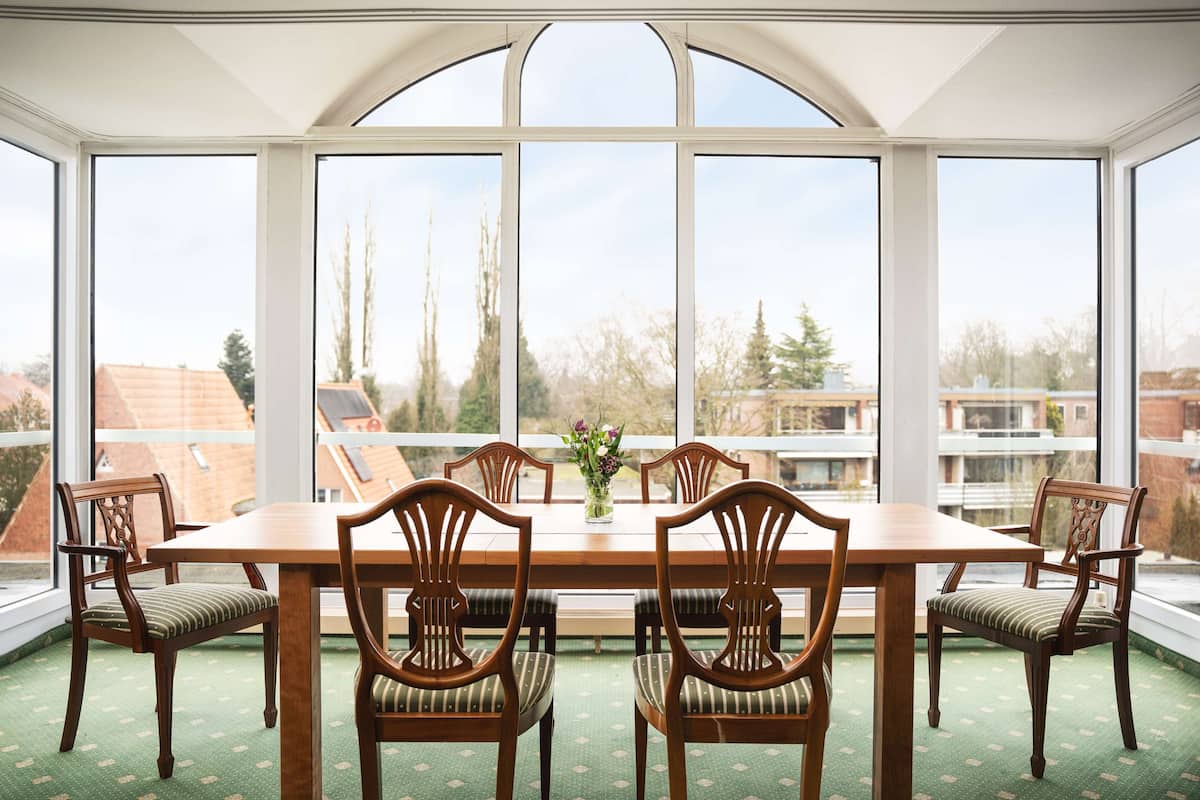 Un comedor con una mesa grande y seis sillas, ventanas amplias con vistas a edificios y árboles, y una alfombra verde estampada.